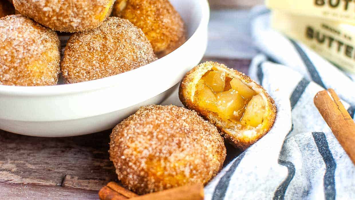A bowl of cinnamon sugar-coated dough balls, one cut open to reveal an apple filling, with butter and cinnamon sticks nearby on a wooden surface.