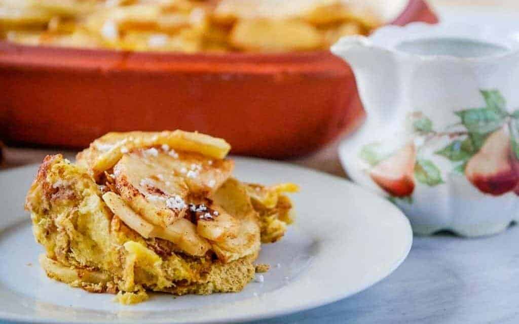 A slice of apple-topped baked French toast sits on a white plate next to a ceramic pitcher with a fruit design.