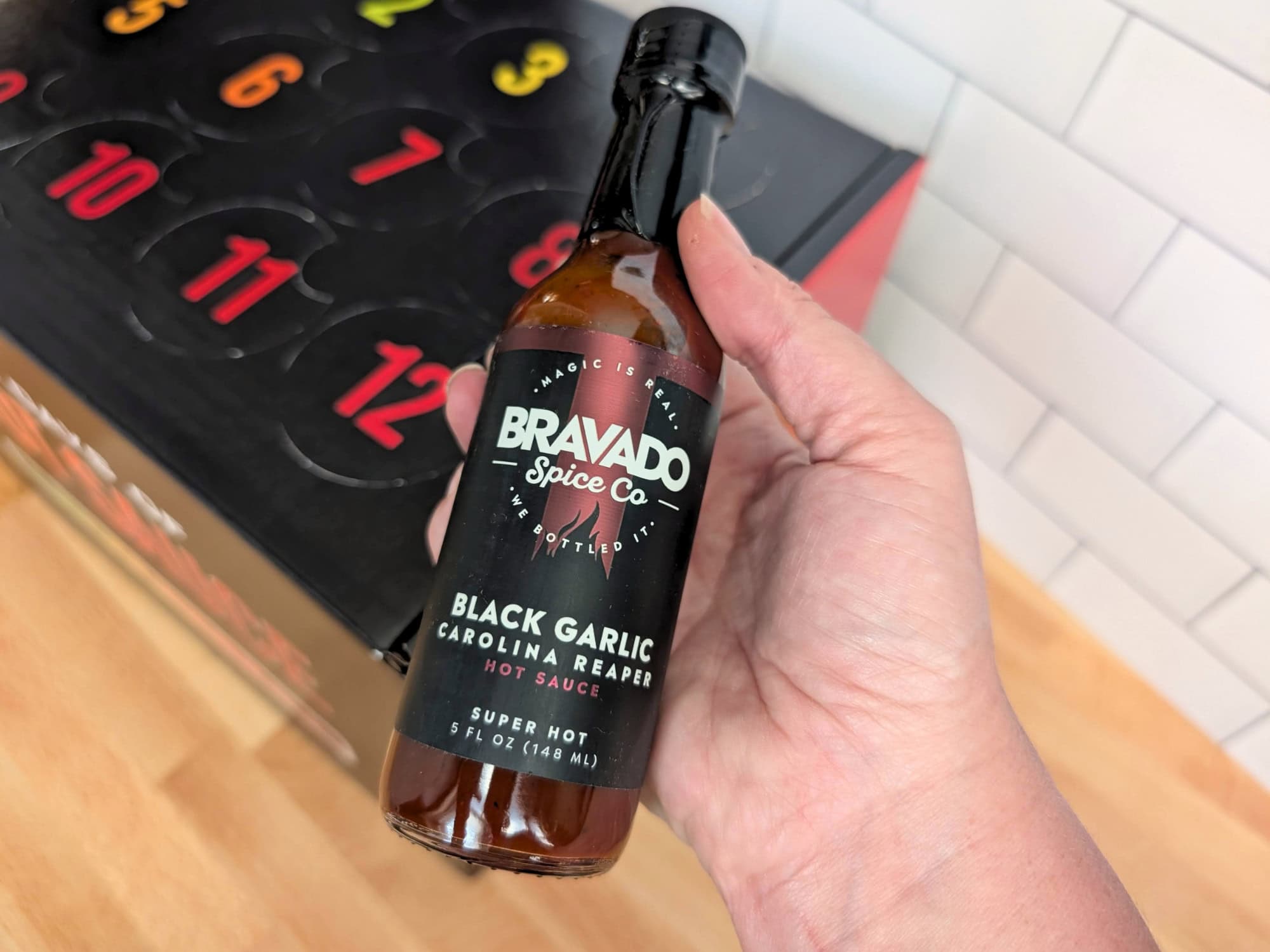 A hand holding a bottle of Bravado Spice Co. Black Garlic Carolina Reaper hot sauce, with a numbered advent calendar in the background.
