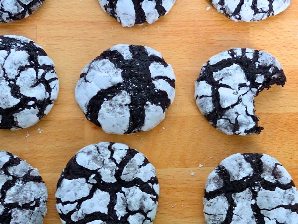 Six chocolate crinkle cookies with powdered sugar on a wooden surface; one cookie has a bite taken out of it.