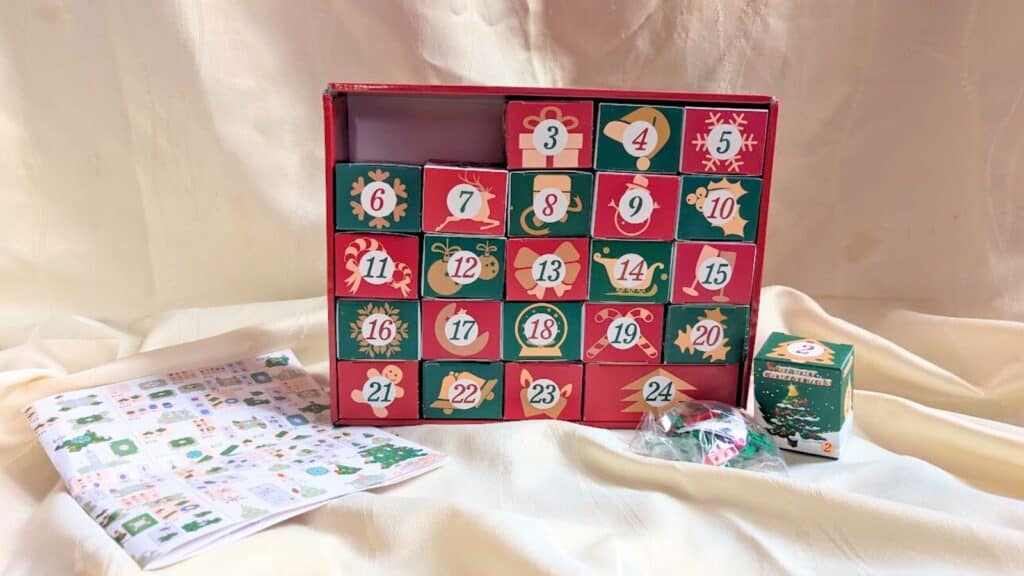 A festive Advent calendar box with numbered drawers, a sheet of stickers, a small decorated box, and a plastic bag with candies displayed in front&mdash;perfect for fans of Advent calendars.