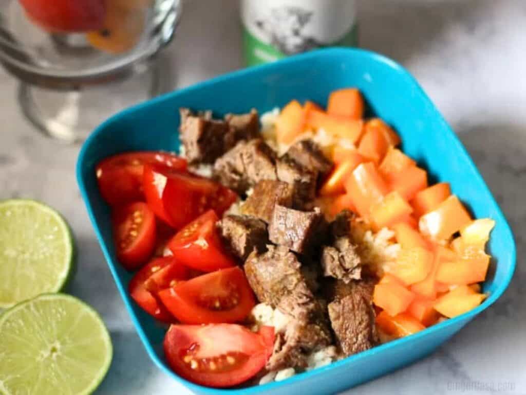 A blue bowl with rice topped with diced beef, halved cherry tomatoes, and chopped orange bell peppers. Lime halves and a glass with fruit are nearby.