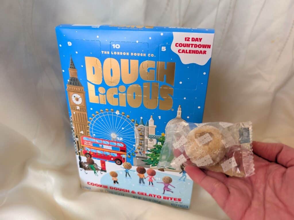 A hand holds an unwrapped cookie dough bite in front of a Doughlicious Advent calendar, designed as a 12-day countdown with charming London-themed illustrations.