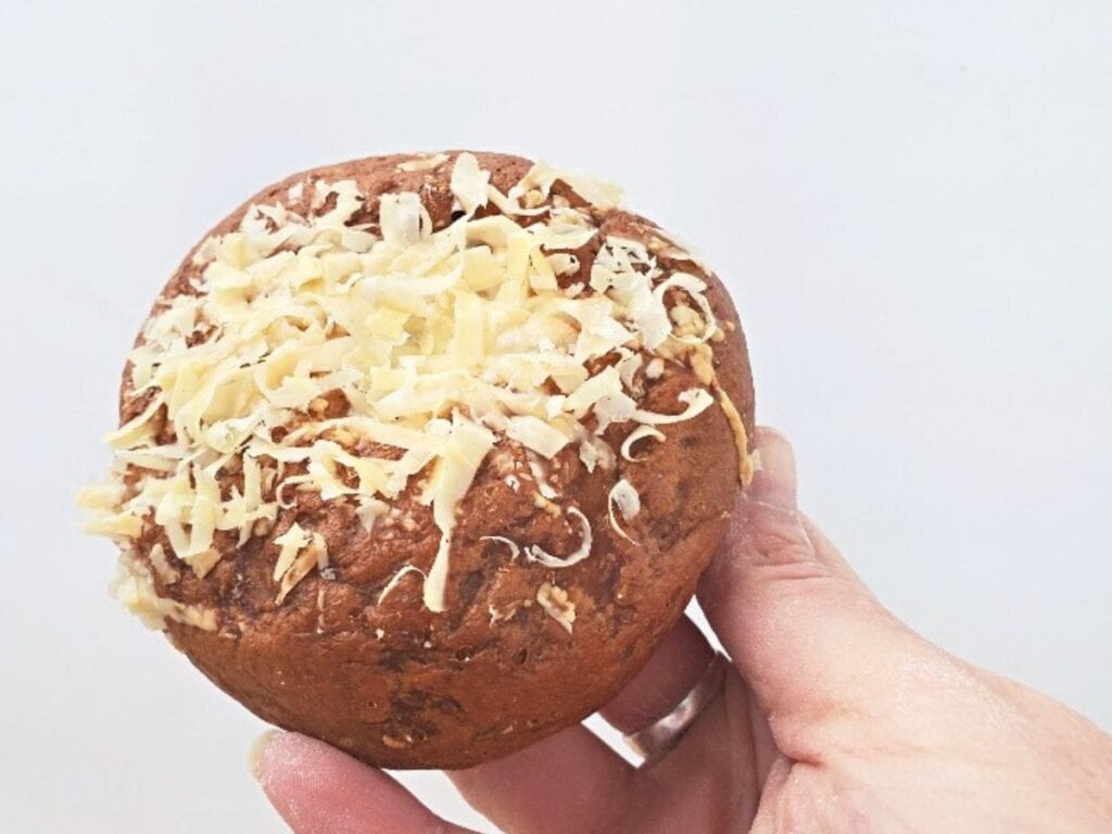 A hand holding a round bread roll topped with shredded cheese against a plain white background.