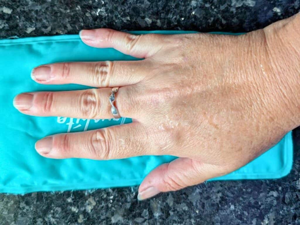 A hand with a silver ring rests on a turquoise ice pack atop a dark granite surface.