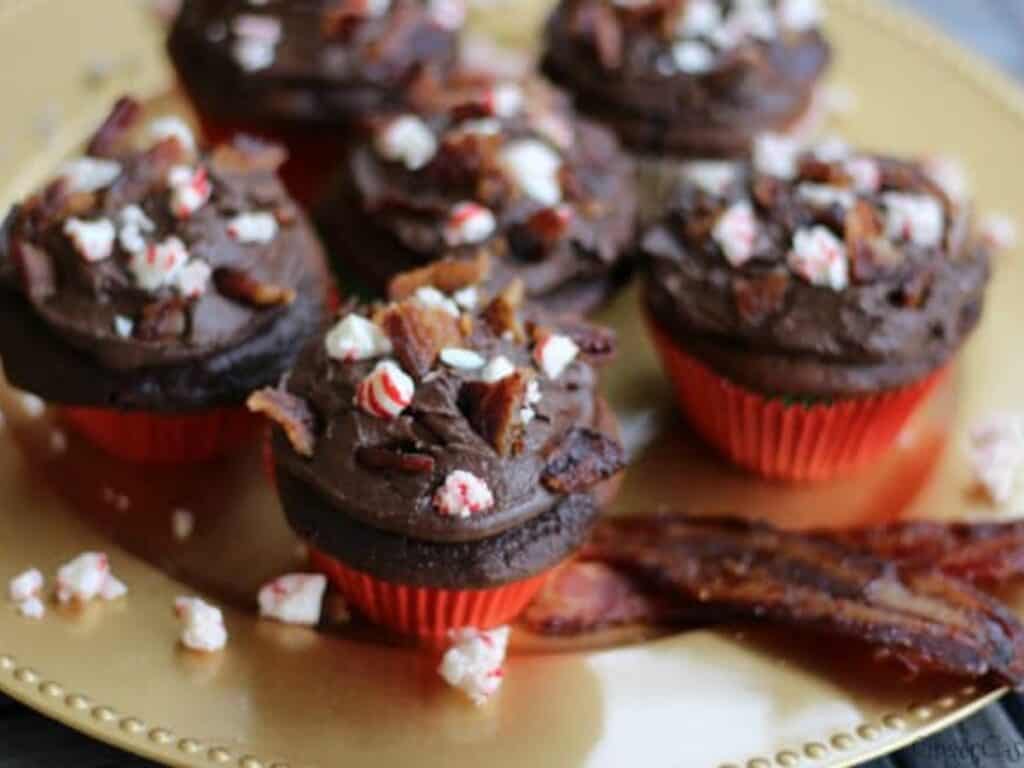 Chocolate cupcakes with dark frosting topped with crumbled candy cane and bacon pieces are arranged on a gold plate with strips of bacon on the side.