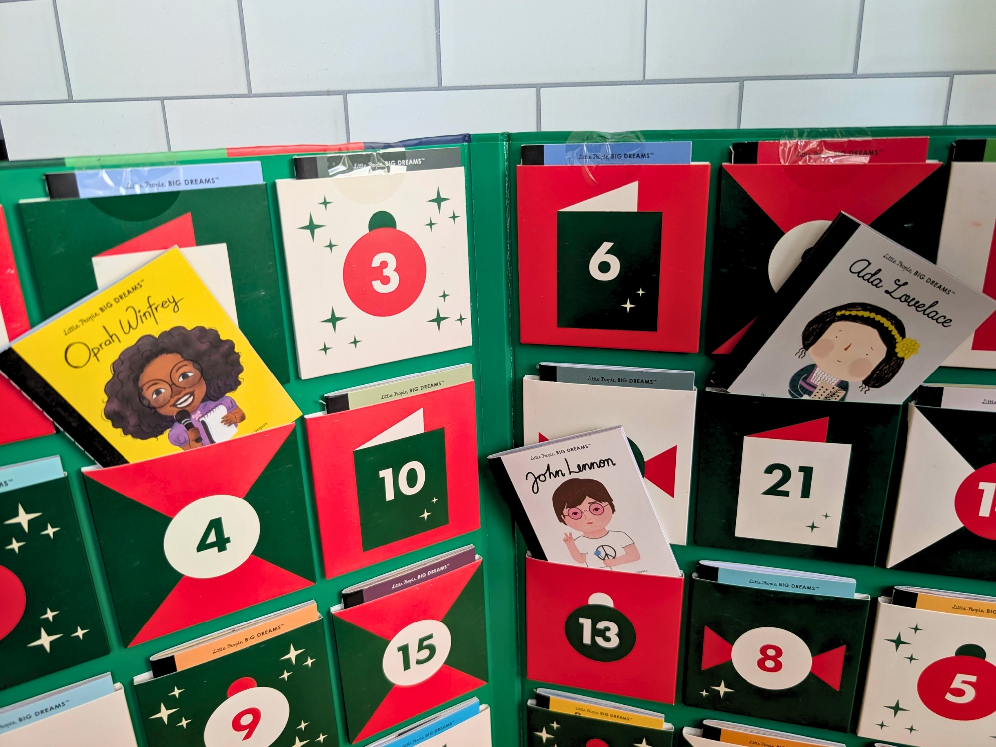 An advent calendar with numbered pockets containing Little People, BIG DREAMS books featuring Oprah Winfrey, John Lennon, and Ada Lovelace.