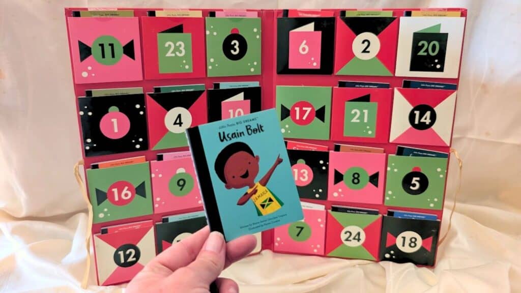 A hand holds a card labeled "Usain Bolt" in front of Advent calendars with numbered doors in shades of pink, green, red, and black.