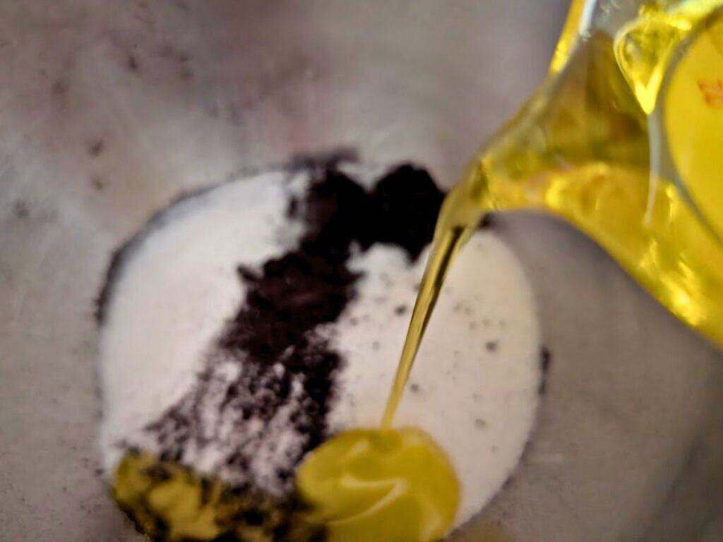Oil is being poured into a metal bowl containing sugar, cocoa powder, and what appears to be butter or margarine.