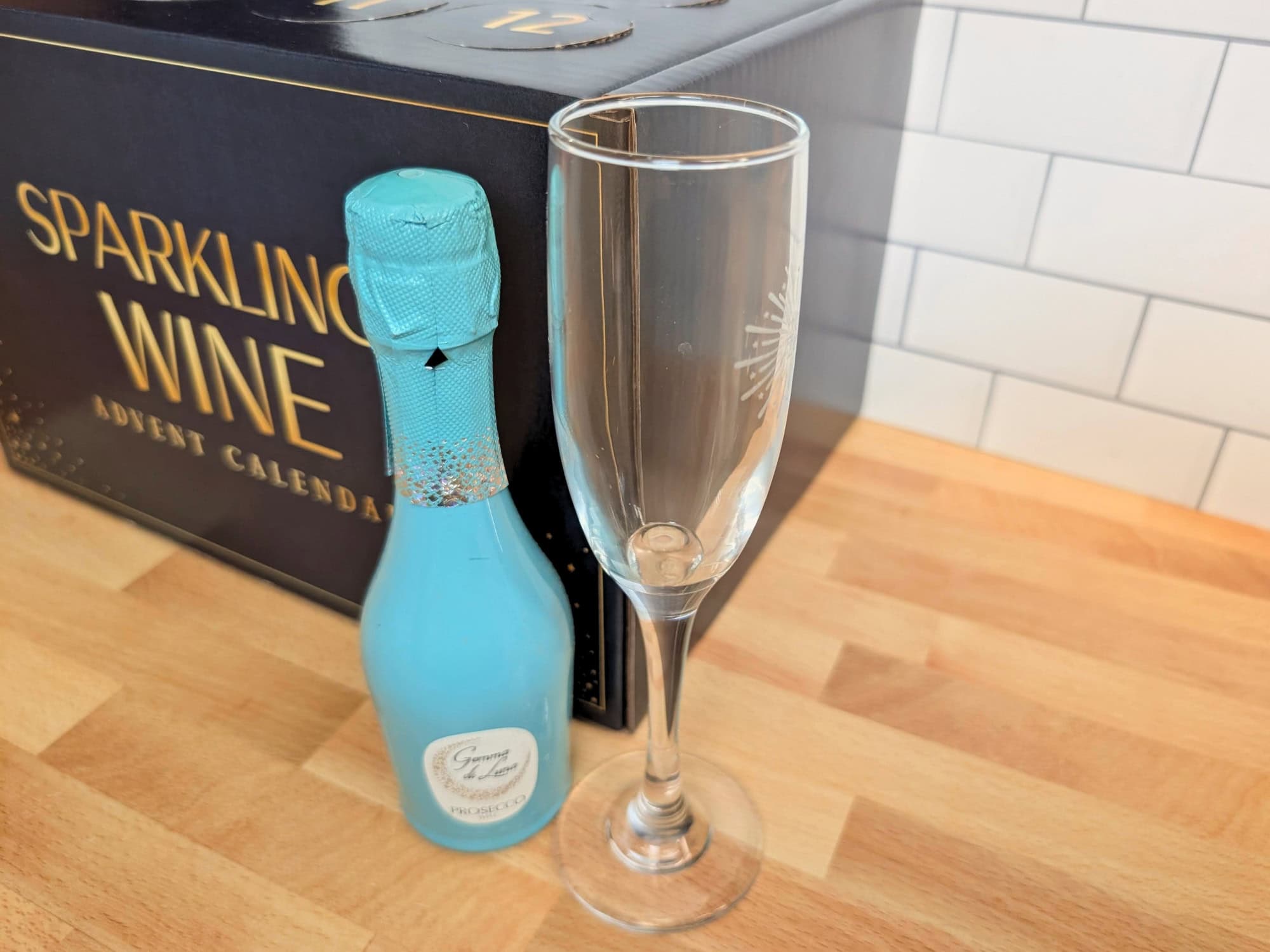 A mini bottle of blue-labeled sparkling wine next to a champagne flute, positioned in front of a Sparkling Wine advent calendar.