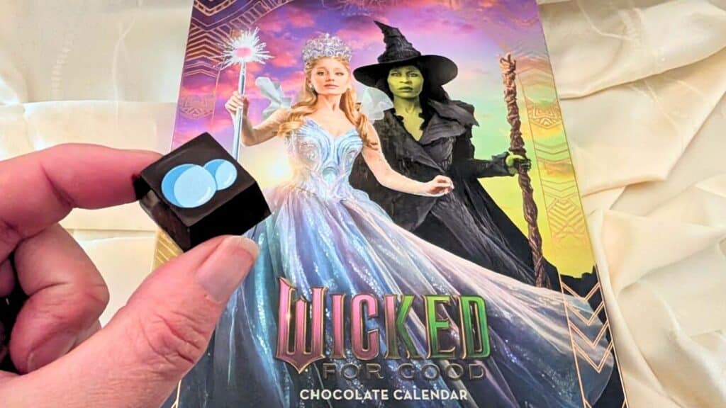 A hand holds a chocolate with a blue design in front of a "Wicked For Good" Advent calendar featuring Glinda and Elphaba from the musical Wicked.