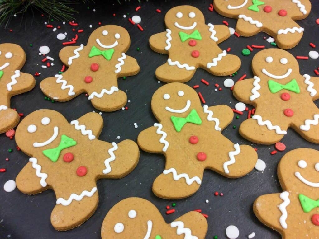 Gingerbread cookies decorated with white, green, and red icing are arranged on a dark surface with festive sprinkles scattered around.