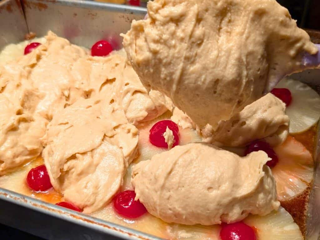 Cake batter being spread over a layer of pineapple rings and maraschino cherries in a metal baking pan, preparing a classic pineapple upside down cake.