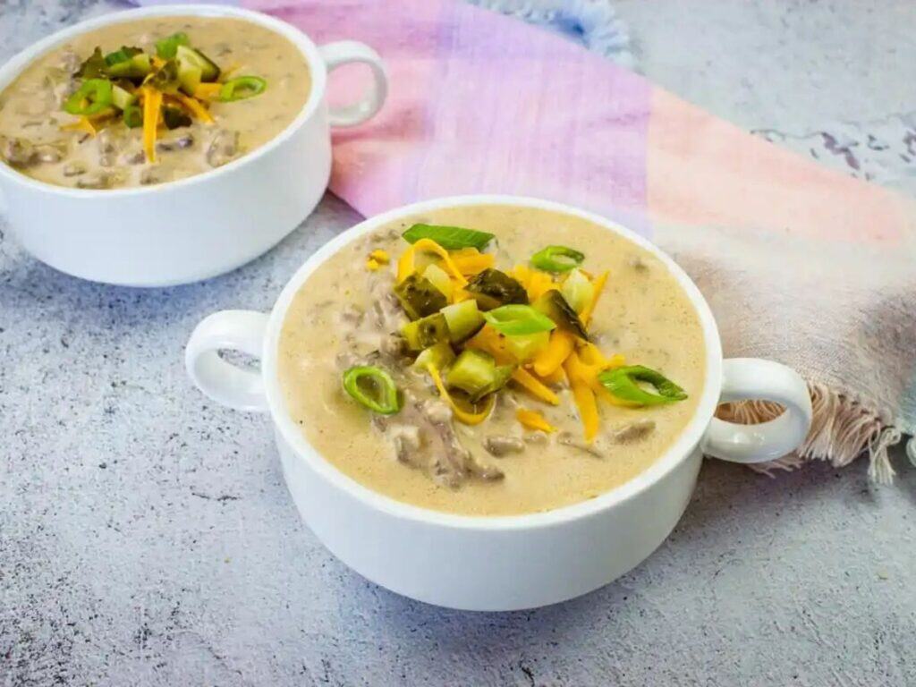 Two white bowls of creamy soup topped with chopped pickles, shredded cheddar cheese, and sliced green onions, placed on a textured surface with a pastel napkin.
