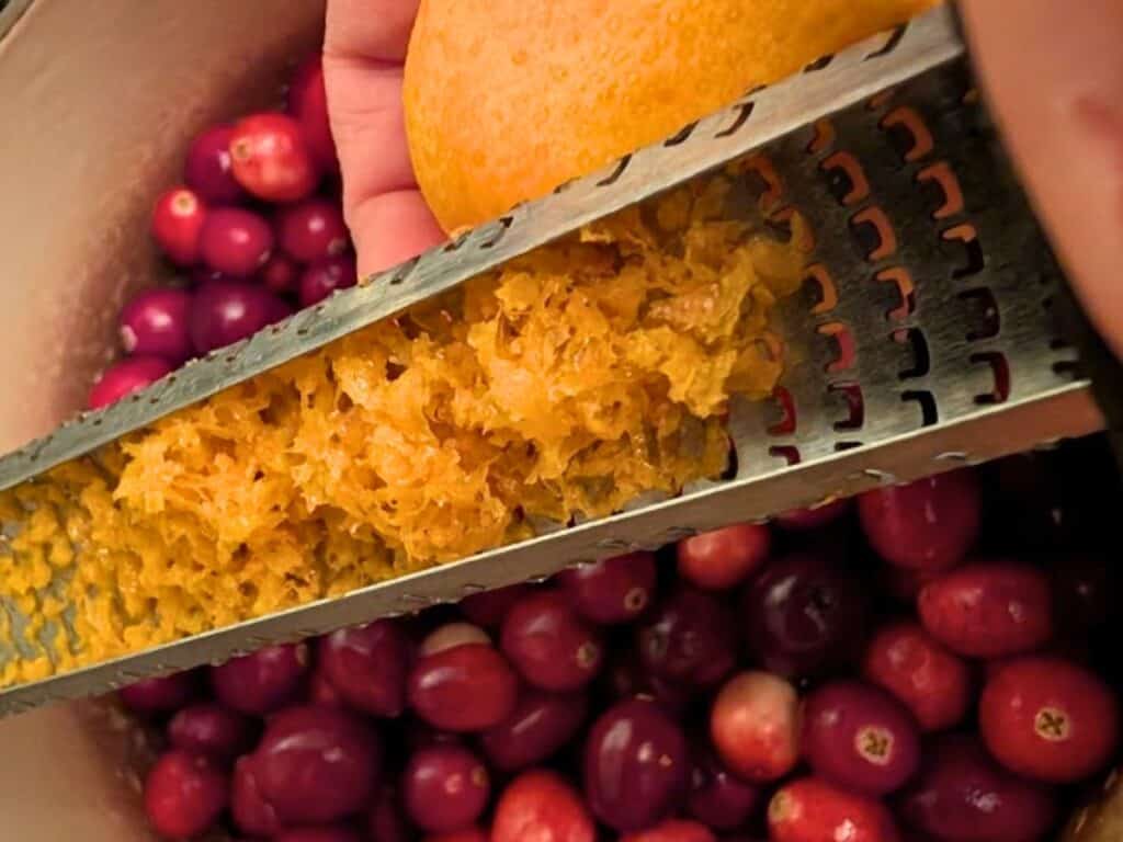 A hand grates orange zest over a bowl filled with fresh cranberries.
