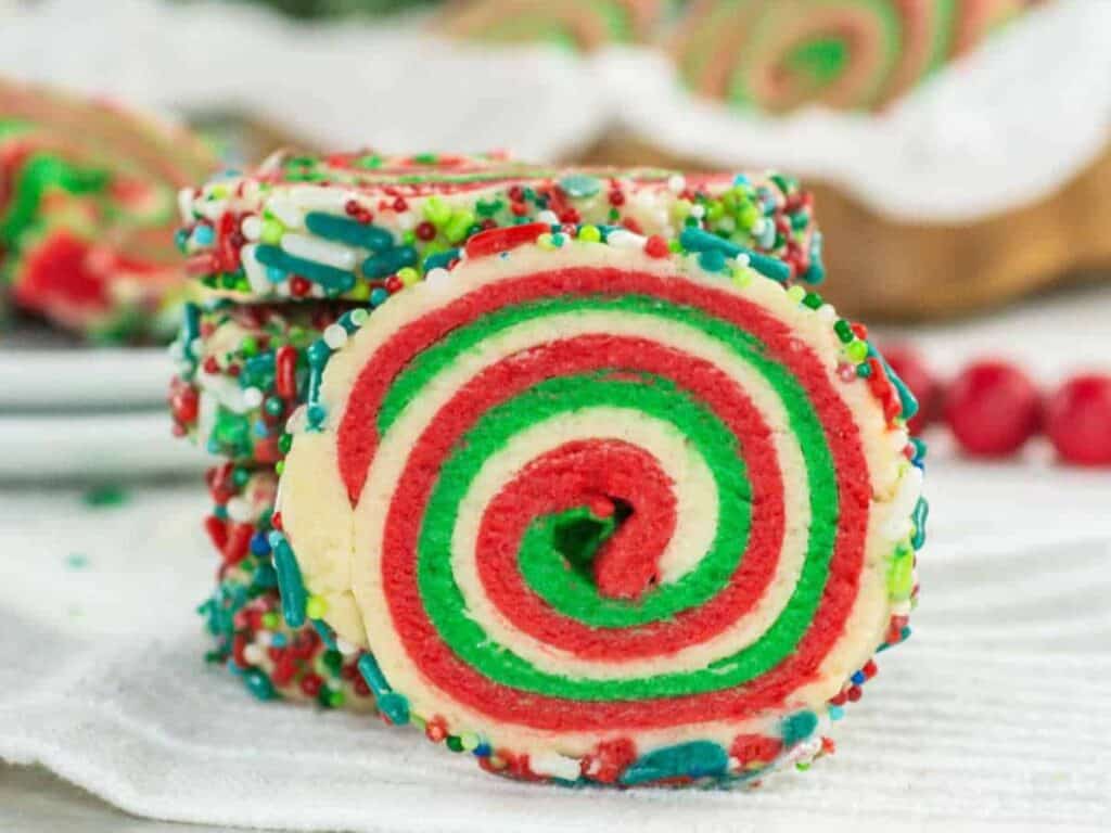 A stack of spiral sugar cookies with red, green, and white swirls, edged with colorful sprinkles, is shown on a white surface.