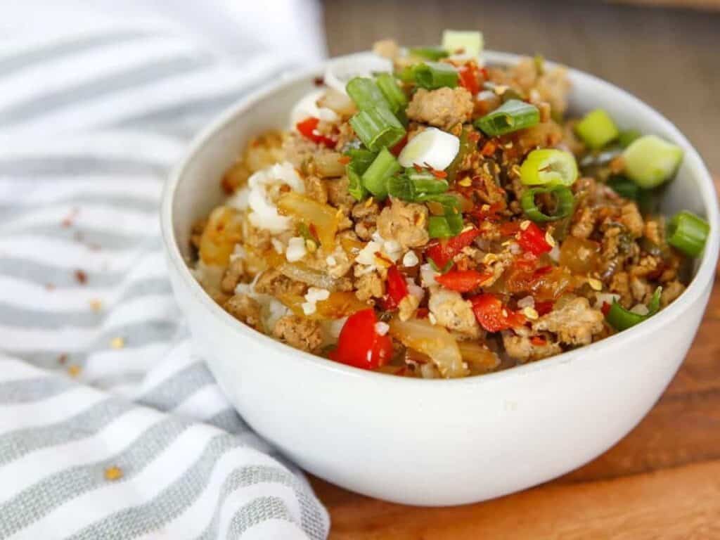 A white bowl filled with a stir-fry featuring ground meat, diced red peppers, onions, and chopped green onions on top, set on a wooden surface next to a striped cloth.
