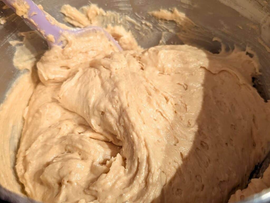 A close-up of thick, light brown pineapple upside down cake batter being mixed in a metal bowl with a purple spatula.