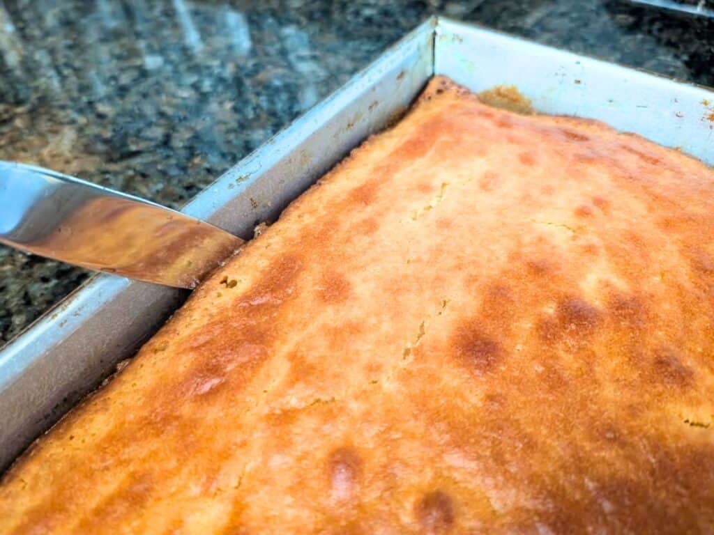 A knife is being used to cut into a rectangular pineapple upside down cake in a metal baking pan on a granite countertop.