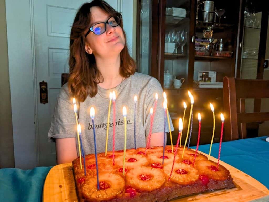 A person wearing glasses sits at a table behind a festive pineapple upside down cake with lit candles.