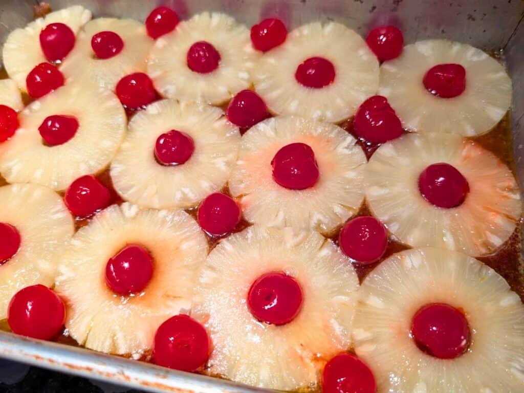 Sliced pineapple rings and red cherries arranged in rows in a metal baking pan, ready to be transformed into a classic pineapple upside down cake.