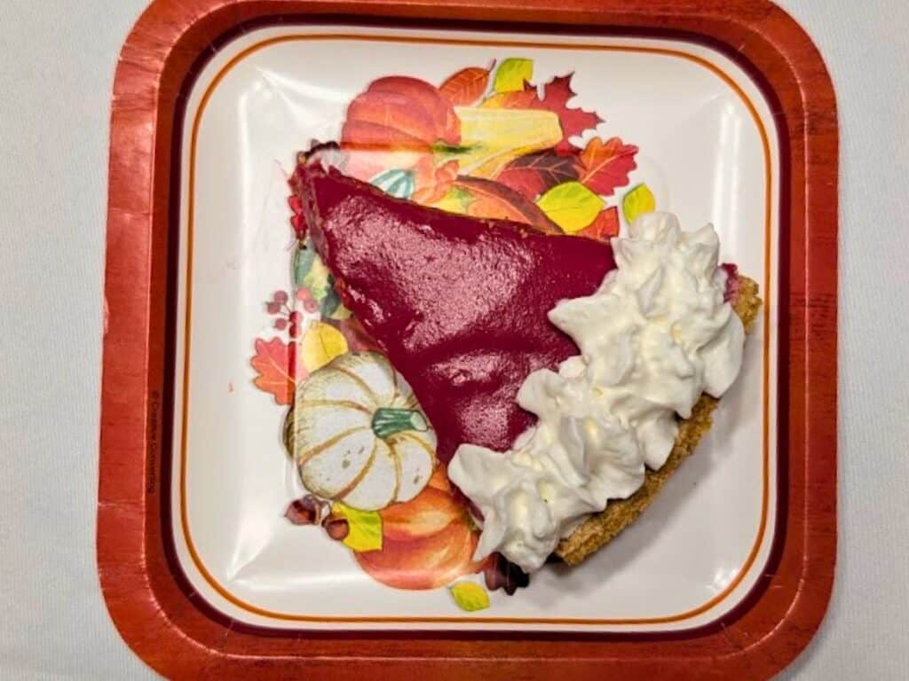 A slice of bright red pie with whipped cream on a fall-themed plate decorated with pumpkins and autumn leaves.
