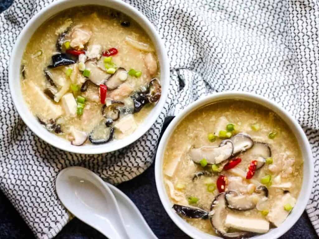 Two bowls of hot and sour soup with mushrooms, tofu, red chili, and green onions, placed on a patterned cloth next to two white ceramic spoons.