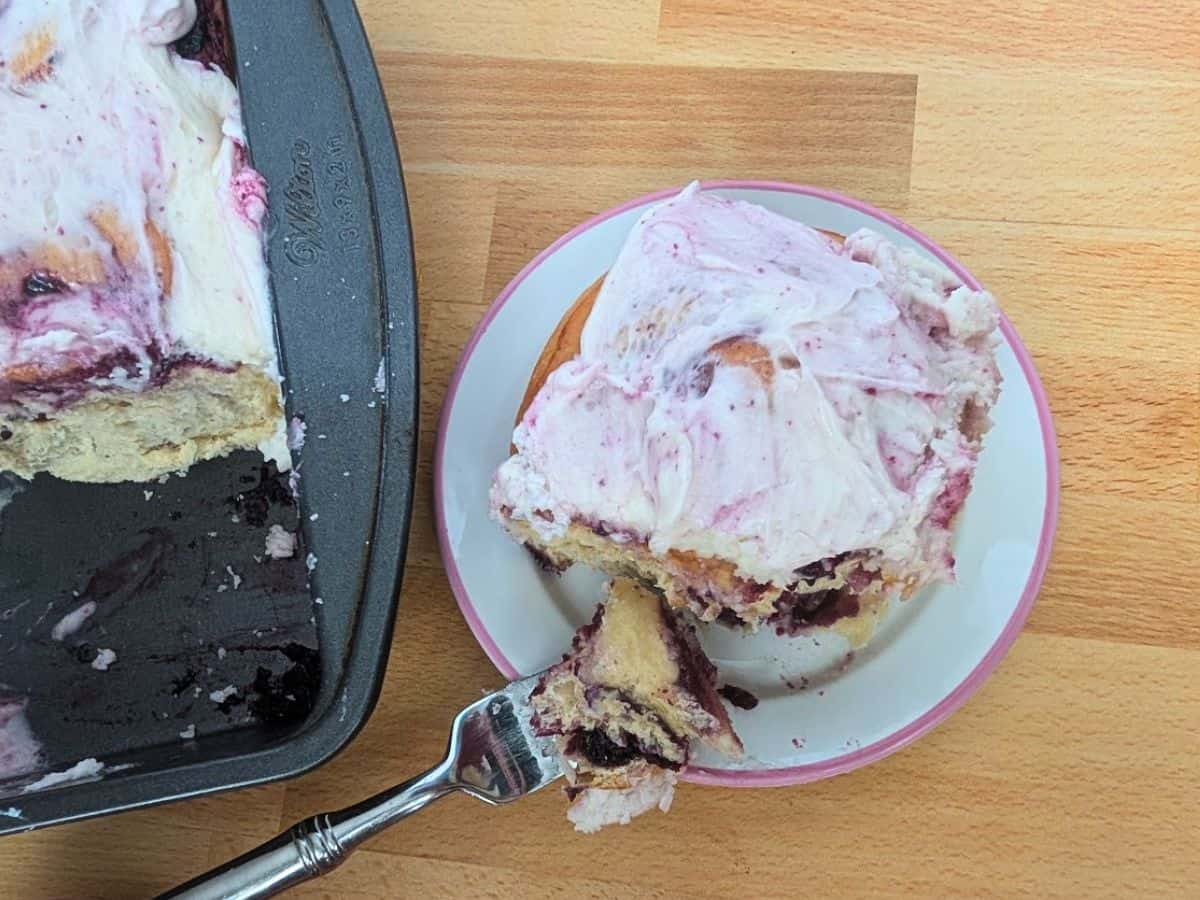 Image shows a plate with a cinnamon roll topped with berry frosting, next to a baking tray with more rolls on a wooden surface. A fork holds a bite-sized piece of the roll.