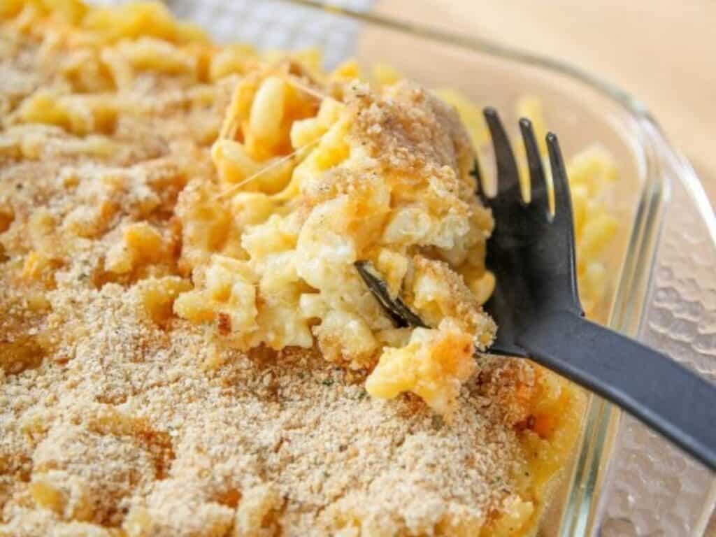 A close-up of baked macaroni and cheese topped with breadcrumbs in a glass dish, with a black plastic fork lifting a portion.