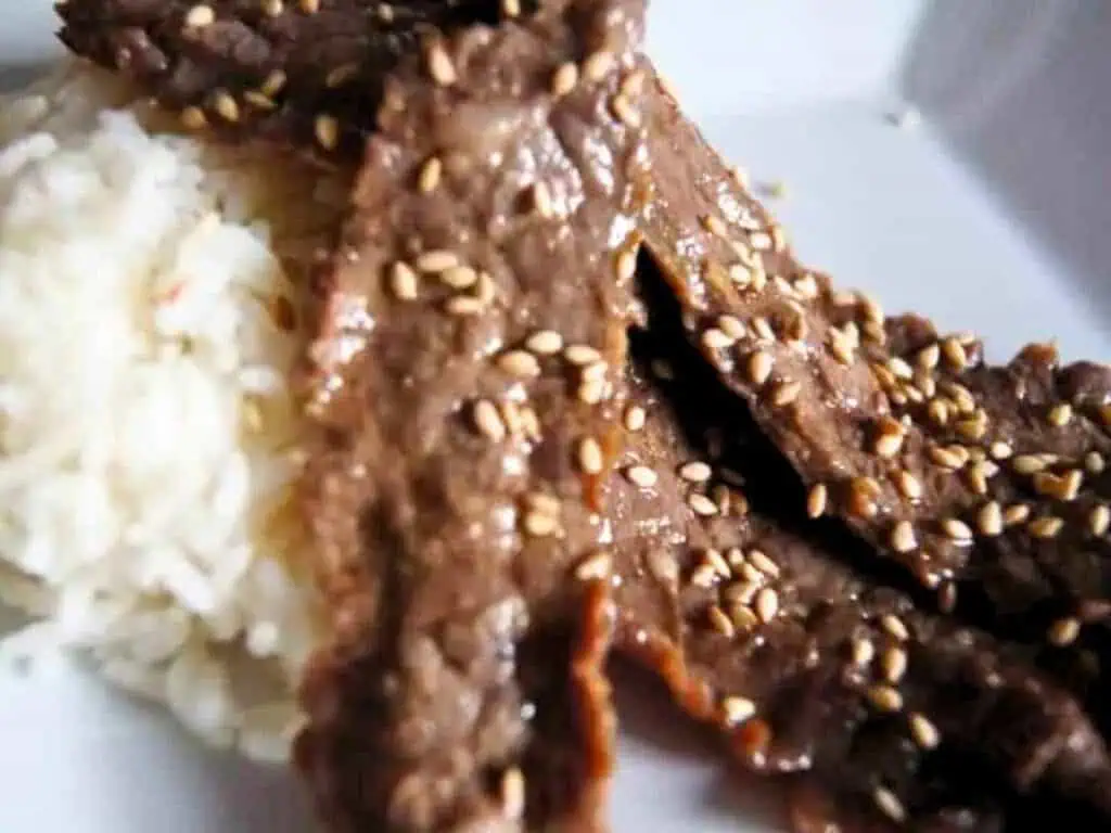 Sliced beef topped with sesame seeds served next to white rice on a plate.