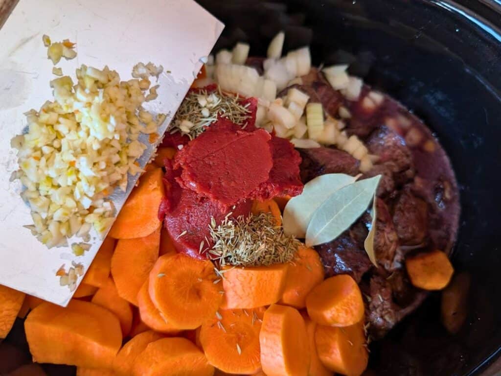 Chopped carrots, onions, garlic, tomato paste, bay leaves, herbs, and beef are assembled in a slow cooker, ready to be cooked.