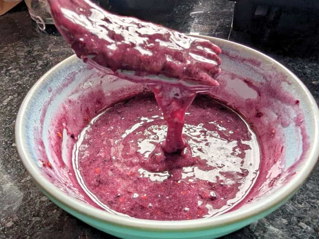A bowl containing a thick, purple batter or mixture, with a utensil lifting some of the batter above the bowl on a dark countertop.