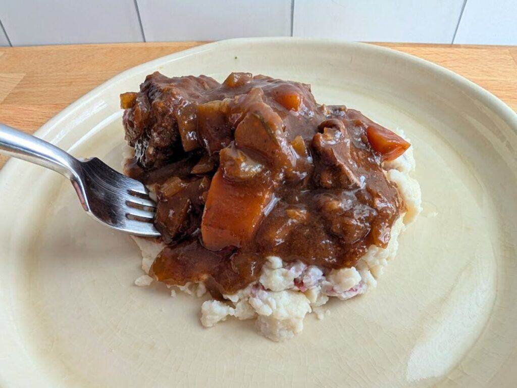 A plate of mashed potatoes topped with beef stew, featuring chunks of beef, carrots, and gravy; a fork is placed on the beef.