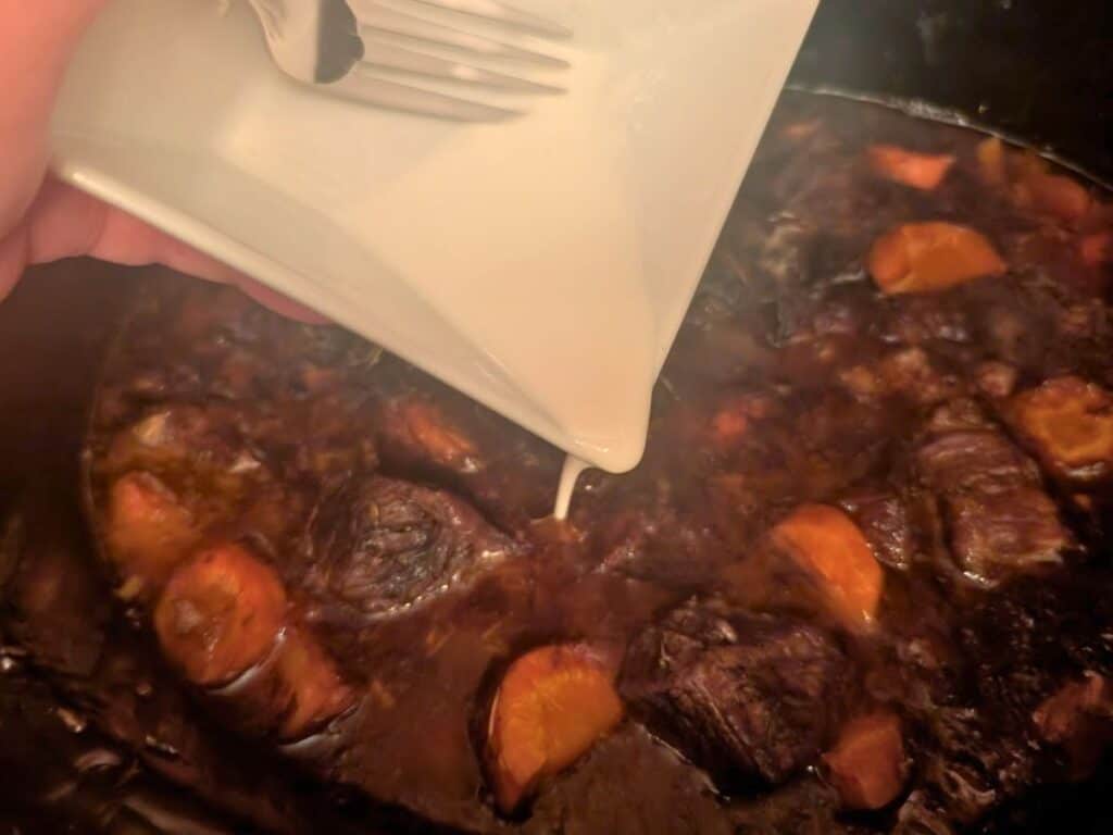 A hand pours liquid from a white bowl into a pot containing chunks of meat and vegetables in a dark sauce.