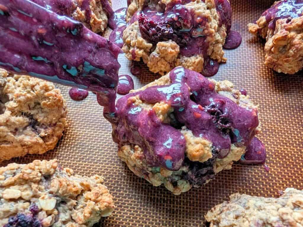 Oatmeal scones with berries on a baking sheet, with purple berry glaze being drizzled over one of the scones.