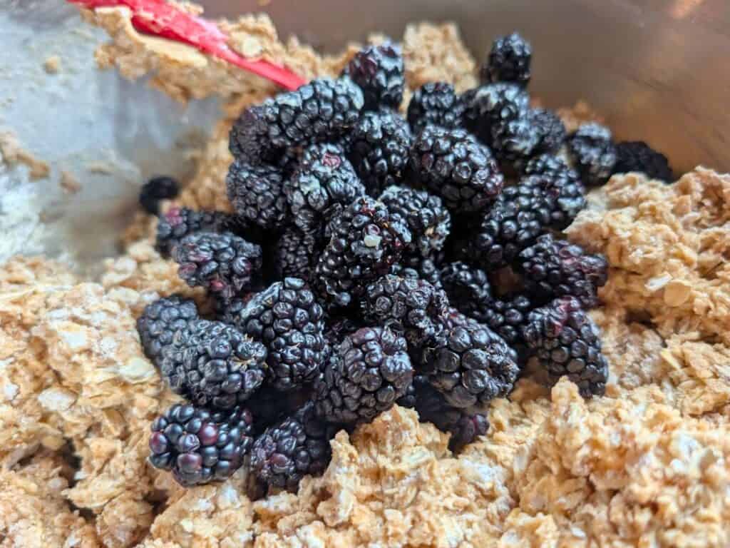 A pile of fresh blackberries sits on top of oatmeal cookie dough in a mixing bowl with a red spatula.
