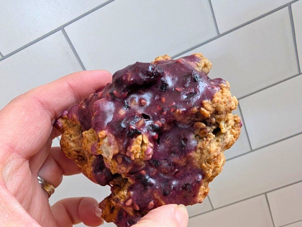 A hand holds an oat cookie topped with a thick layer of purple berry glaze in front of a white tiled wall.