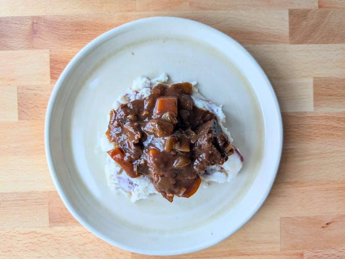 A serving of beef stew with carrots and onions over mashed potatoes on a white plate, set on a wooden surface.