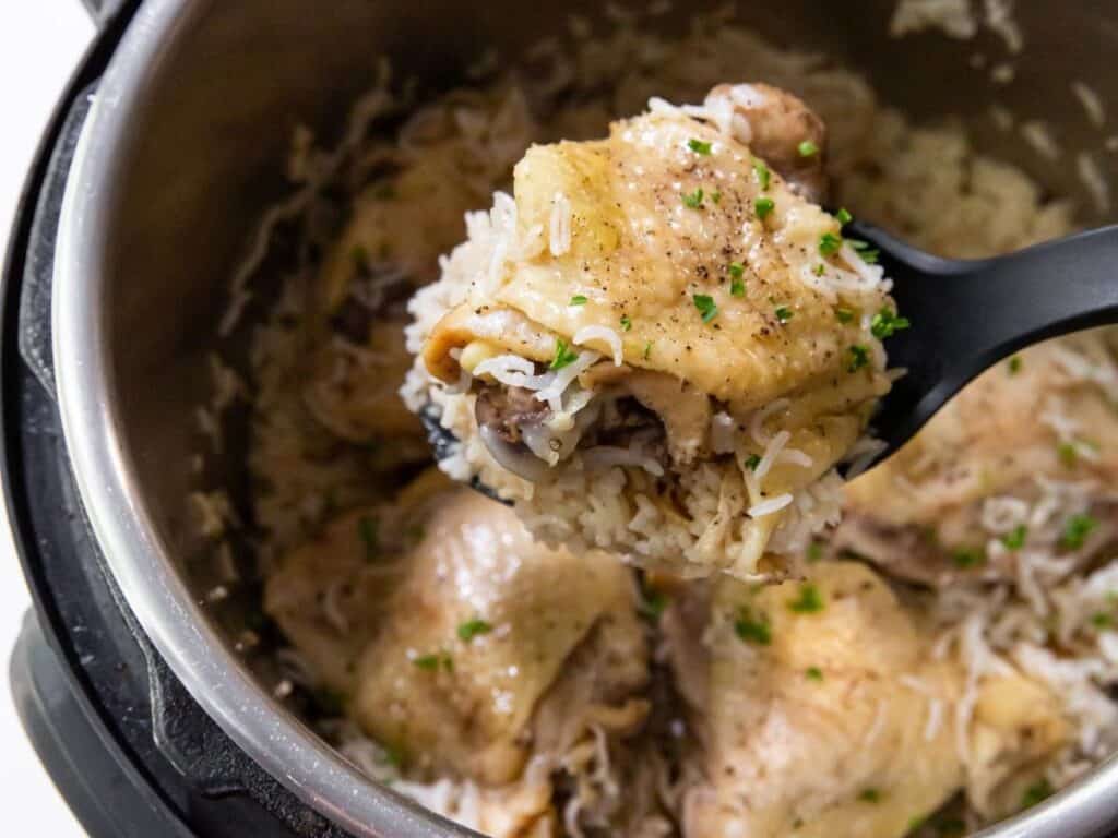 A serving spoon lifts a portion of cooked chicken and rice from an Instant Pot. The dish is garnished with chopped herbs.