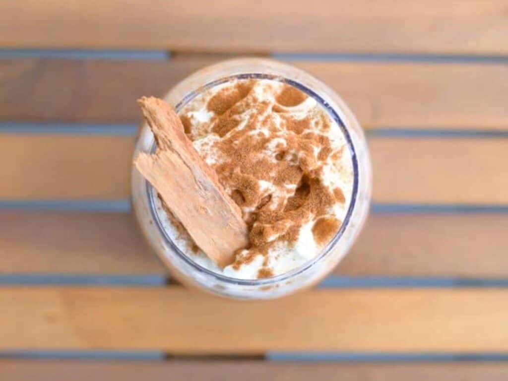 A glass of creamy drink topped with whipped cream, ground cinnamon, and a stick of cinnamon, placed on a wooden slatted surface.
