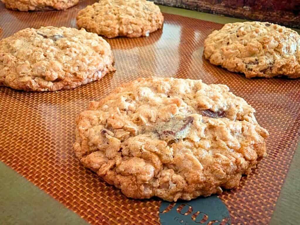Several large, round oatmeal cookies with chocolate chunks rest on a silicone baking mat.