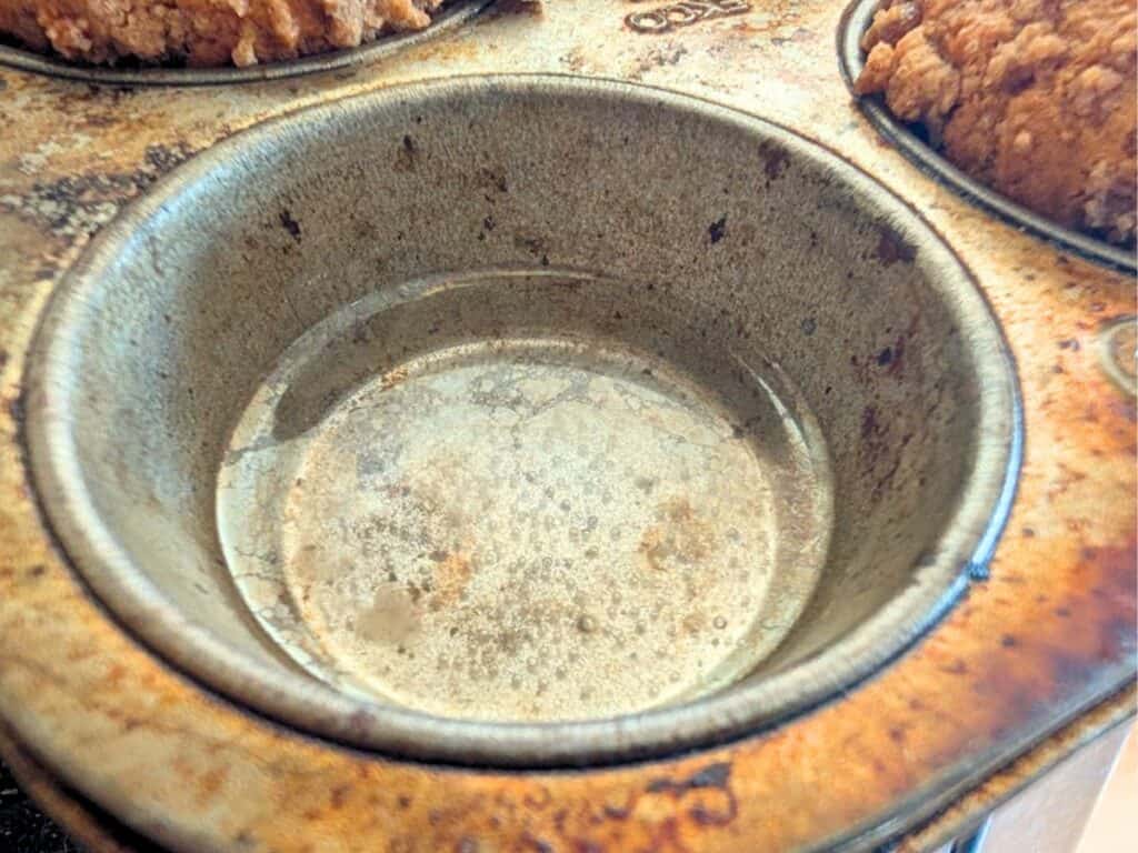 An empty muffin cup filled halfway with water next to baked pumpkin streusel muffins in the same tin.