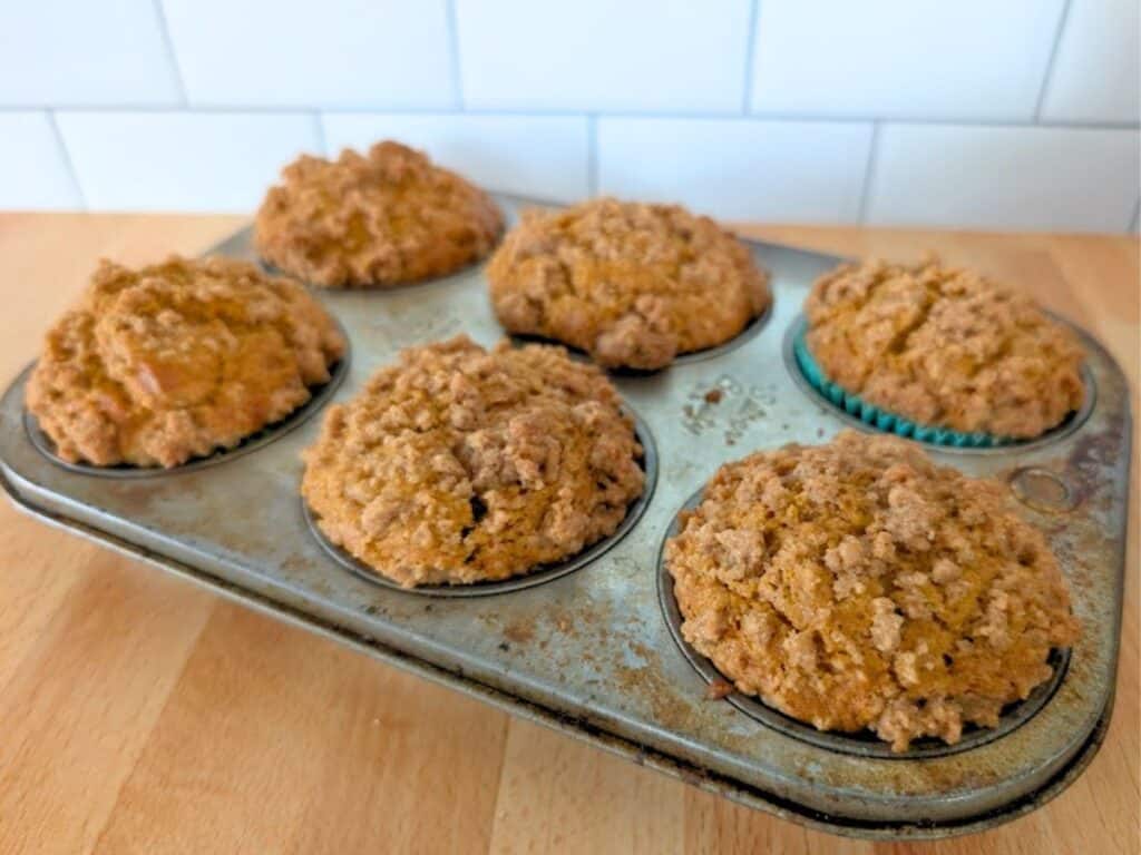 Six baked muffins with a crumb topping sit in a metal muffin tin on a wooden surface in front of a white tiled wall.