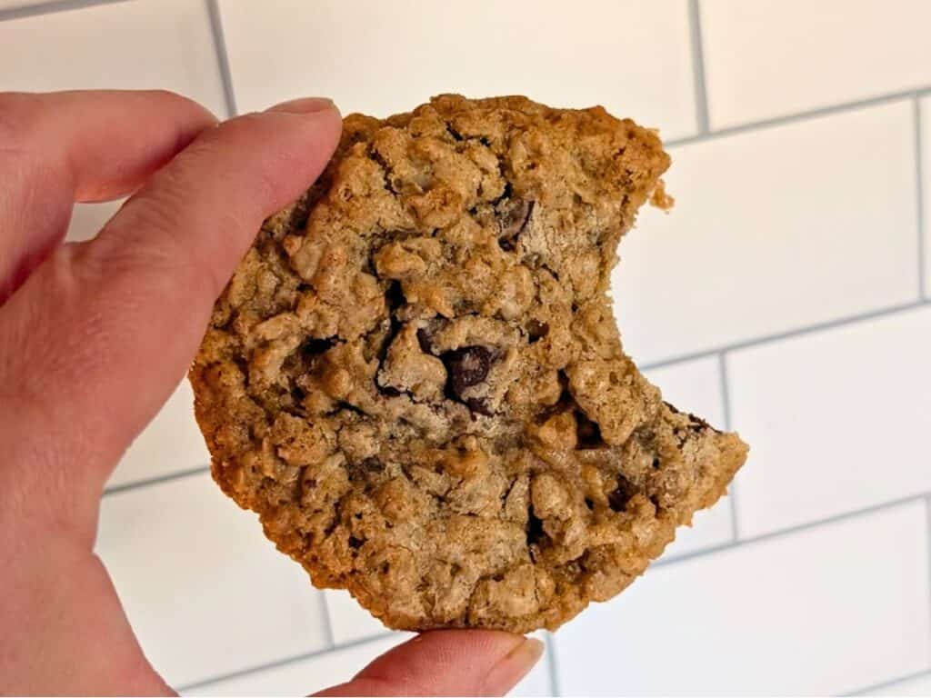 A hand holding an oatmeal chocolate chip cookie with a bite taken out of it, against a white tiled background.