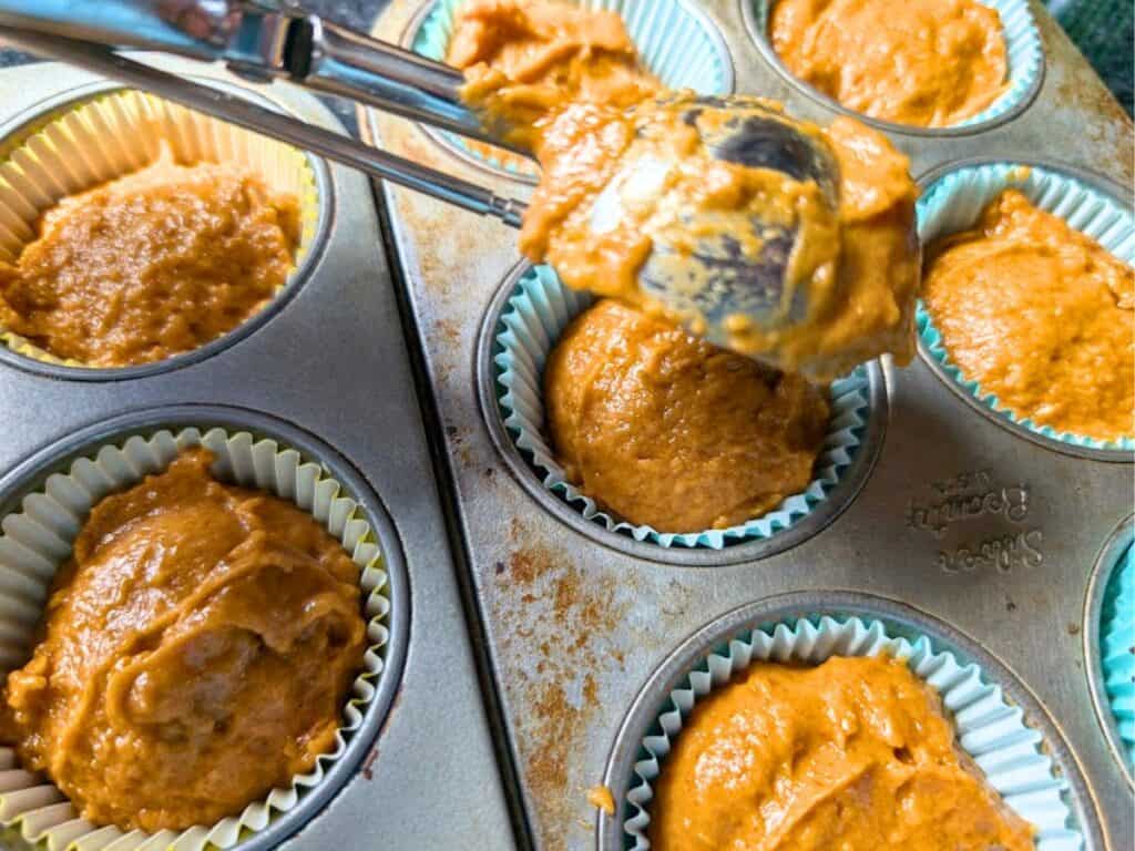 Close-up of a muffin tin lined with paper cups and filled with orange batter, as a scoop is used to fill one of the cups.