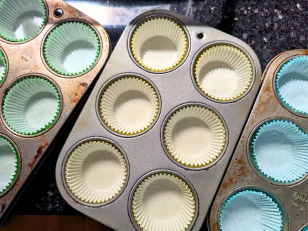Three muffin pans with empty paper liners are arranged on a granite countertop, ready for baking.