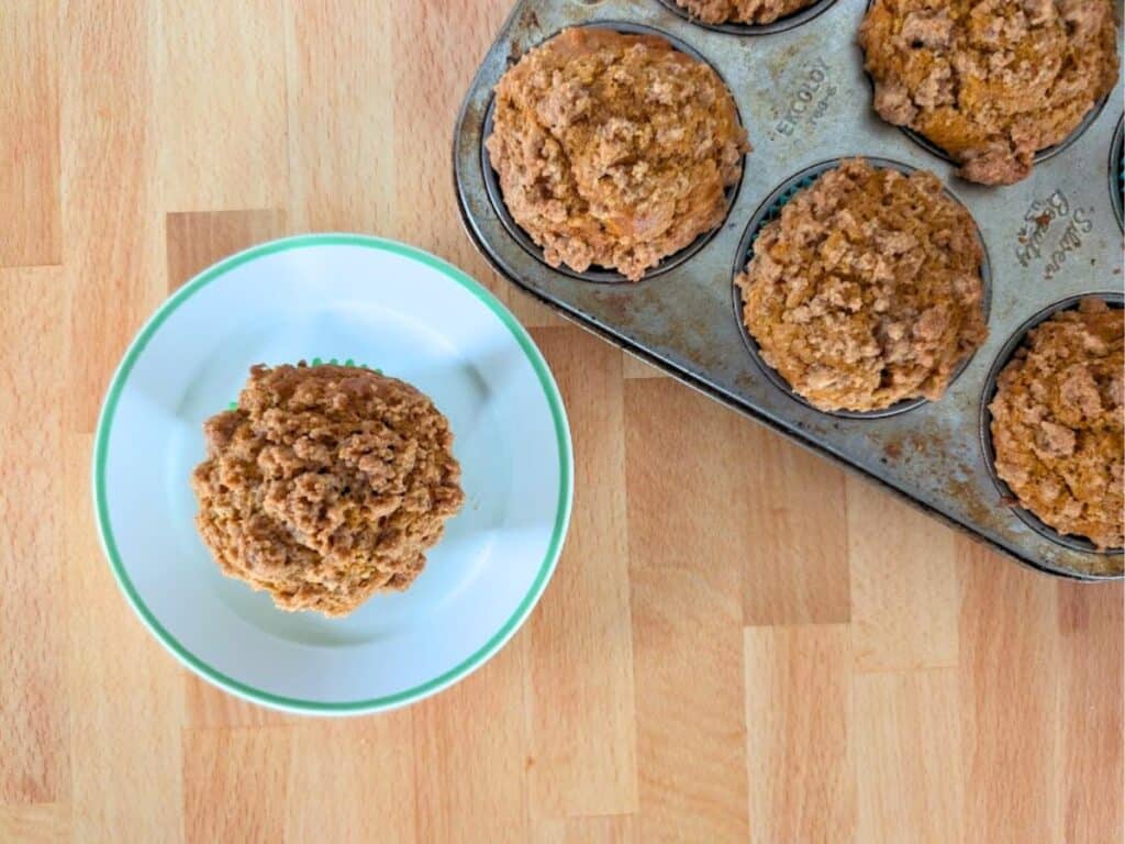 A plate with a single muffin sits next to a metal muffin tin holding five more muffins on a wooden surface.