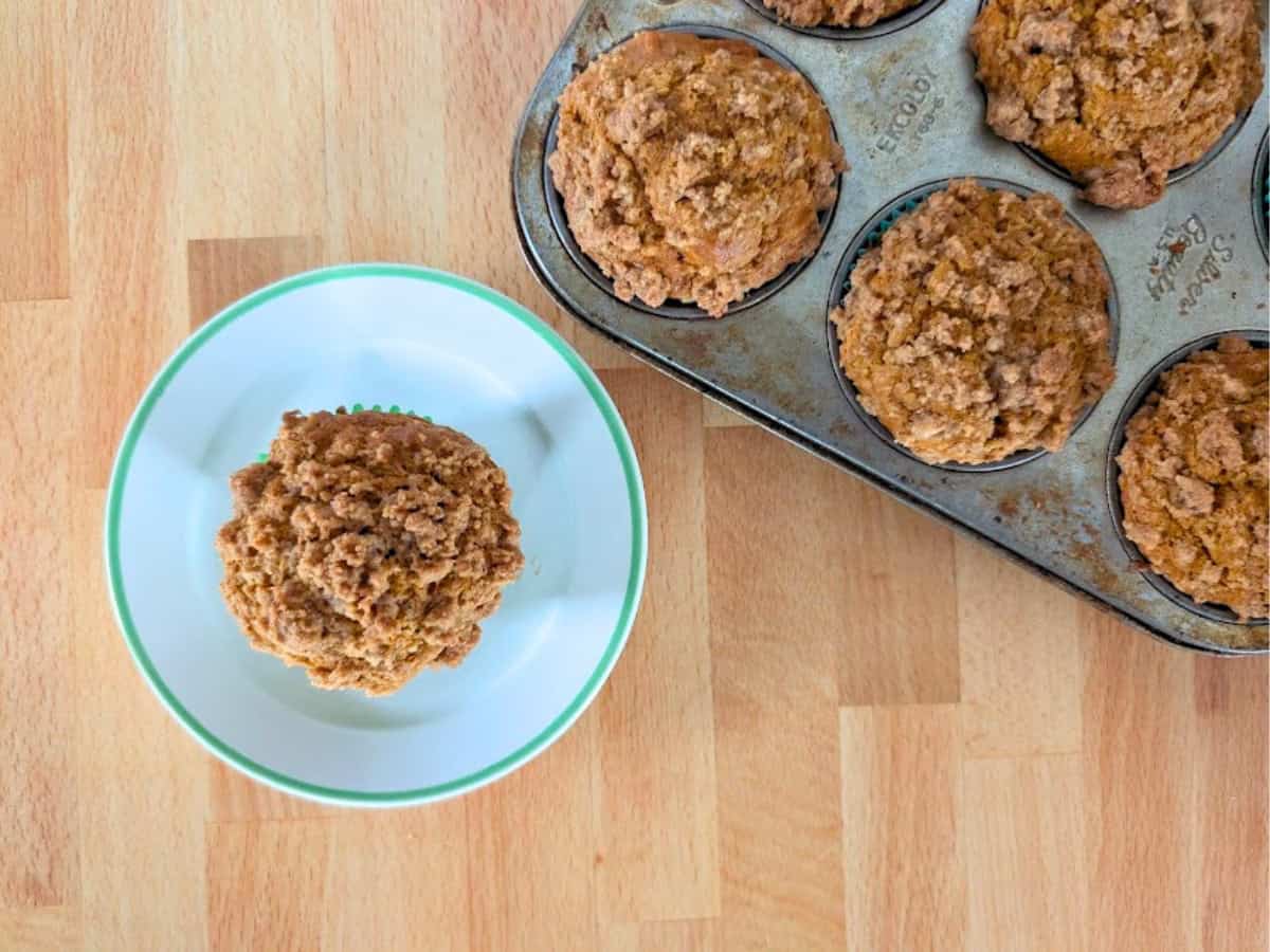 A plate with a single muffin sits next to a metal muffin tin holding five more muffins on a wooden surface.