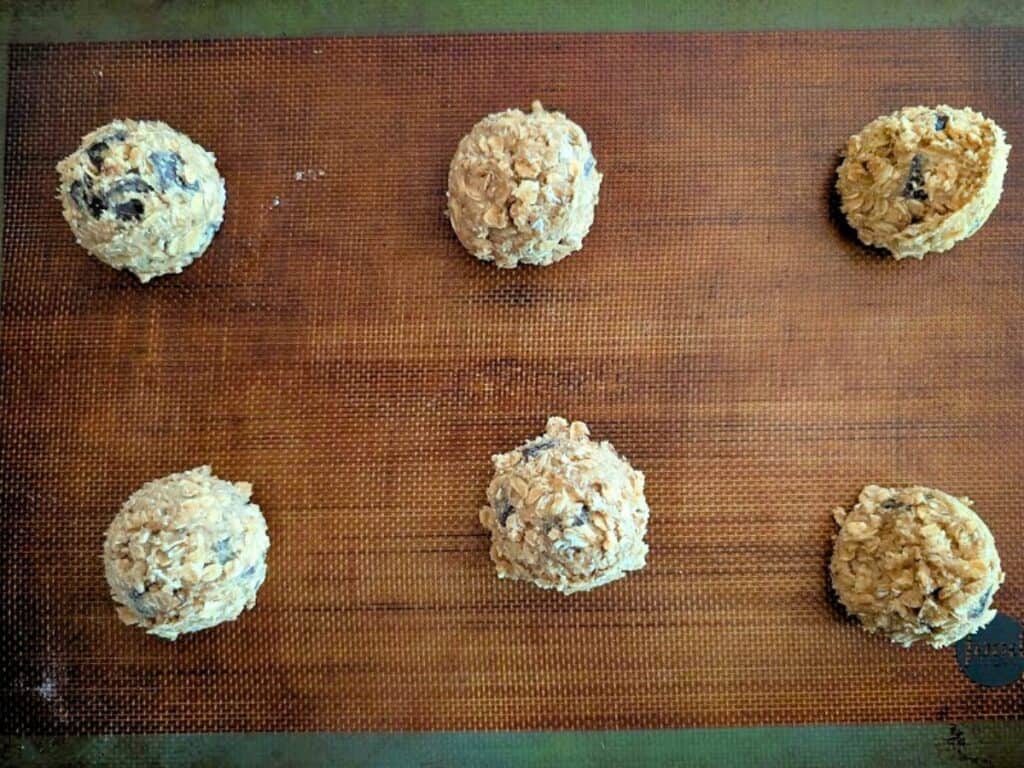 Six scoops of oatmeal cookie dough with chocolate chips spaced evenly on a brown silicone baking mat, ready to be baked.
