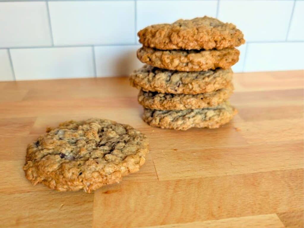 Five oatmeal cookies are stacked on a wooden surface, with one cookie lying flat beside the stack. White subway tile backsplash in the background.